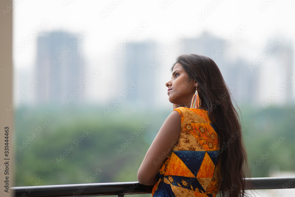 Fashion portrait of an young and beautiful brunette dark skinned Bengali woman in vibrant indo-western dress standing on a balcony in green urban background. Indian lifestyle.