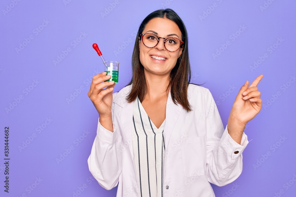 Young beautiful brunette scientist woman wearing coat and glasses holding test tube Smiling happy pointing with hand and finger to the side Young beautiful brunette scientist woman wearing coat and glasses holding test tube Smiling happy pointing with hand and finger to the side