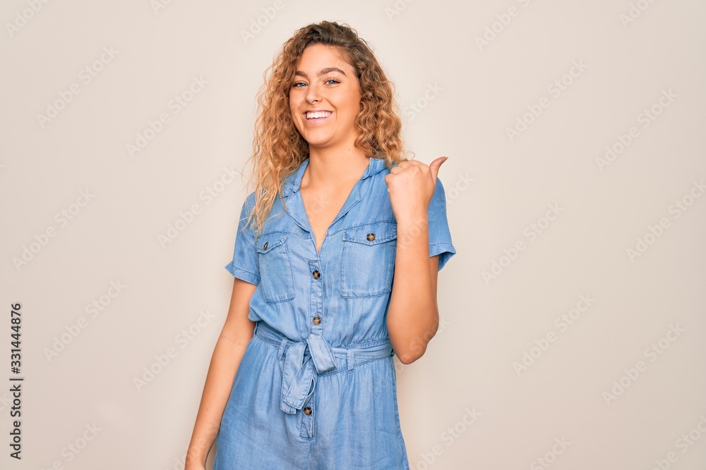 Young beautiful woman with blue eyes wearing casual denim dress over white background smiling with happy face looking and pointing to the side with thumb up. Young beautiful woman with blue eyes wearing casual denim dress over white background smiling with happy face looking and pointing to the side with thumb up.