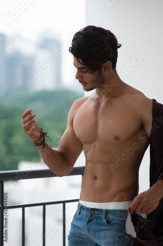 Portrait of an young and handsome brunette Bengali muscular man standing in bare body on a balcony in green urban background. Indian lifestyle. © abir