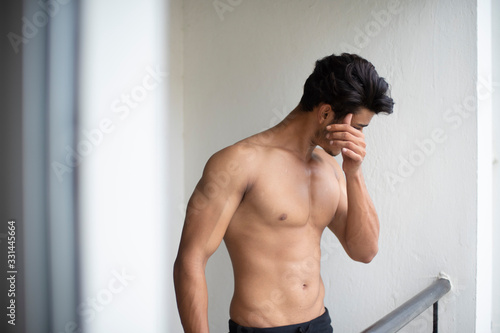 Portrait of an young and handsome brunette Bengali muscular man standing in bare body on a balcony in green urban background. Indian lifestyle.