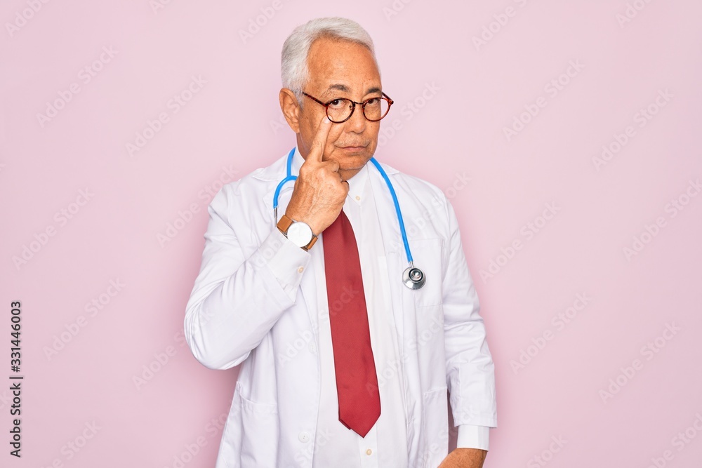Middle age senior grey-haired doctor man wearing stethoscope and professional medical coat Pointing to the eye watching you gesture, suspicious expression Middle age senior grey-haired doctor man wearing stethoscope and professional medical coat Pointing to the eye watching you gesture, suspicious expression