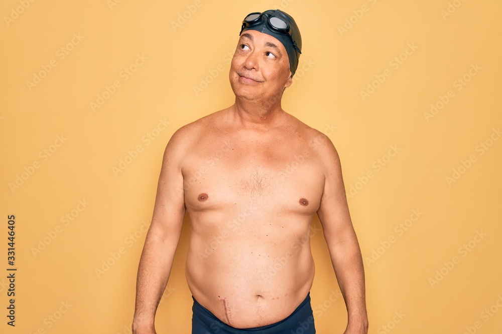 Middle age senior grey-haired swimmer man wearing swimsuit, cap and goggles smiling looking to the side and staring away thinking.