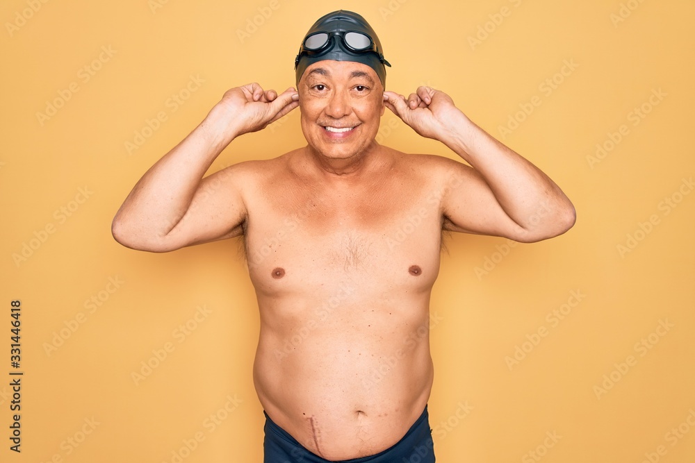 Middle age senior grey-haired swimmer man wearing swimsuit, cap and goggles Smiling pulling ears with fingers, funny gesture. Audition problem