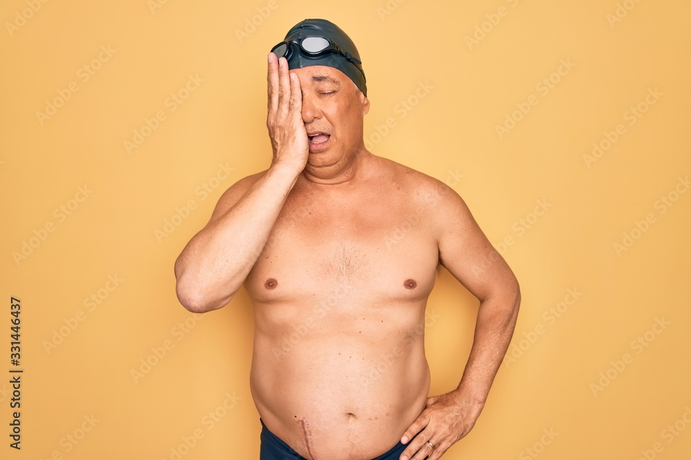 Middle age senior grey-haired swimmer man wearing swimsuit, cap and goggles Yawning tired covering half face, eye and mouth with hand. Face hurts in pain.