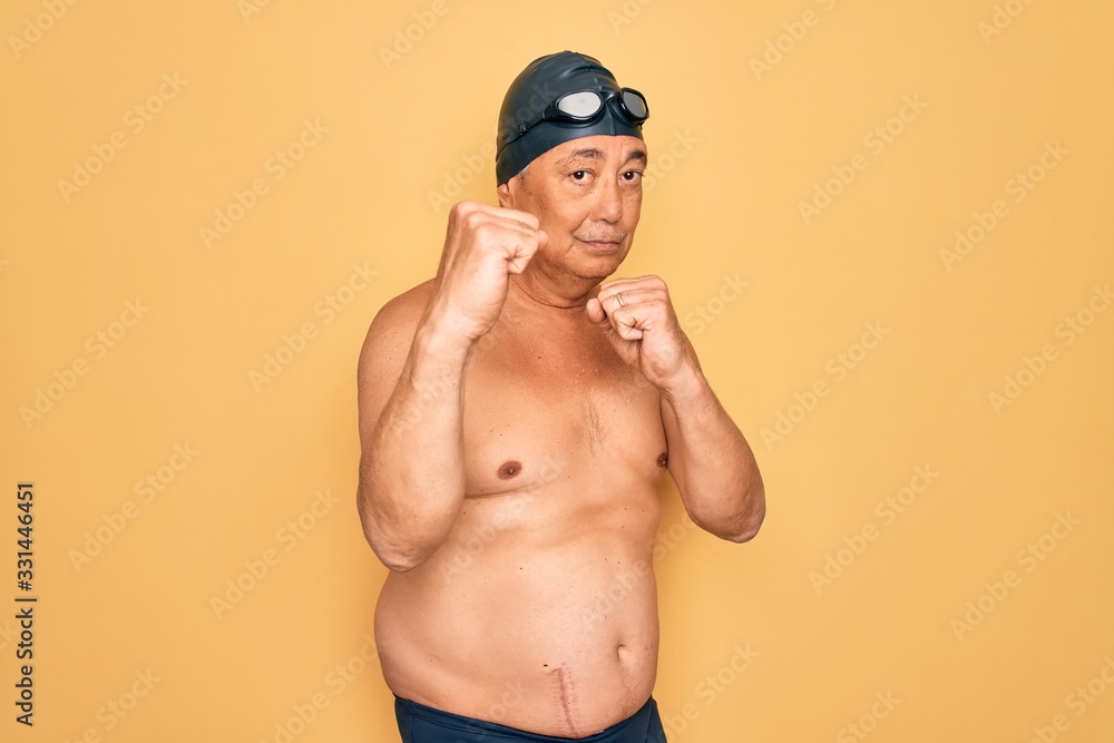 Middle age senior grey-haired swimmer man wearing swimsuit, cap and goggles Ready to fight with fist defense gesture, angry and upset face, afraid of problem