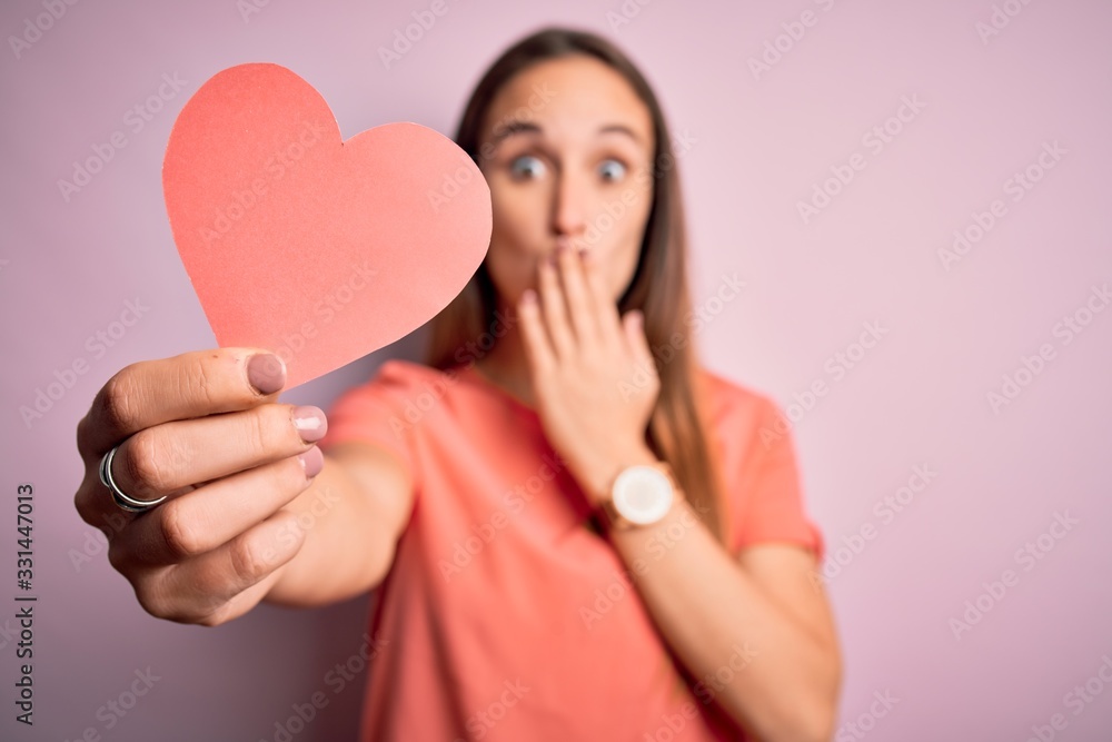 Young beautiful romantic woman holding paper heart shape over isolated pink background cover mouth with hand shocked with shame for mistake, expression of fear, scared in silence, secret concept Young beautiful romantic woman holding paper heart shape over isolated pink background cover mouth with hand shocked with shame for mistake, expression of fear, scared in silence, secret concept