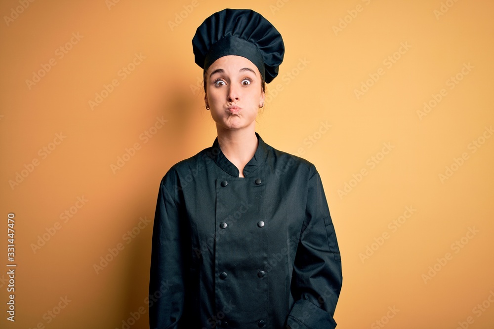 Young beautiful chef woman wearing cooker uniform and hat standing over yellow background puffing cheeks with funny face. Mouth inflated with air, crazy expression. Young beautiful chef woman wearing cooker uniform and hat standing over yellow background puffing cheeks with funny face. Mouth inflated with air, crazy expression.