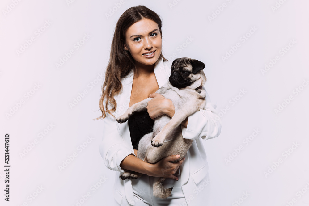 A laughing and smiling auburn haired woman in a white dress, is staring most lovingly at her cute pug, who calmly sits on the hands, gaining her undivided attention. Isolated white background.