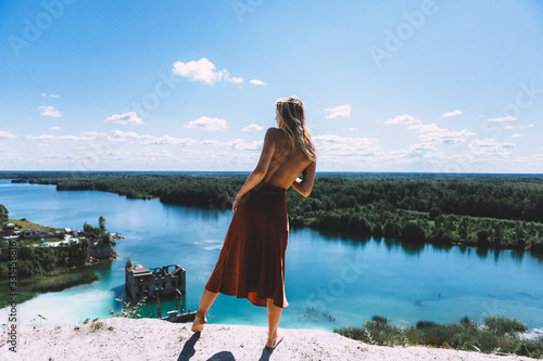 At a picturesque vantage point of Rummu quarry, a blond woman is standing in her bare feet, wearing a thigh high, auburn coloured dress, With her back turned away from the camera. Clear Water.