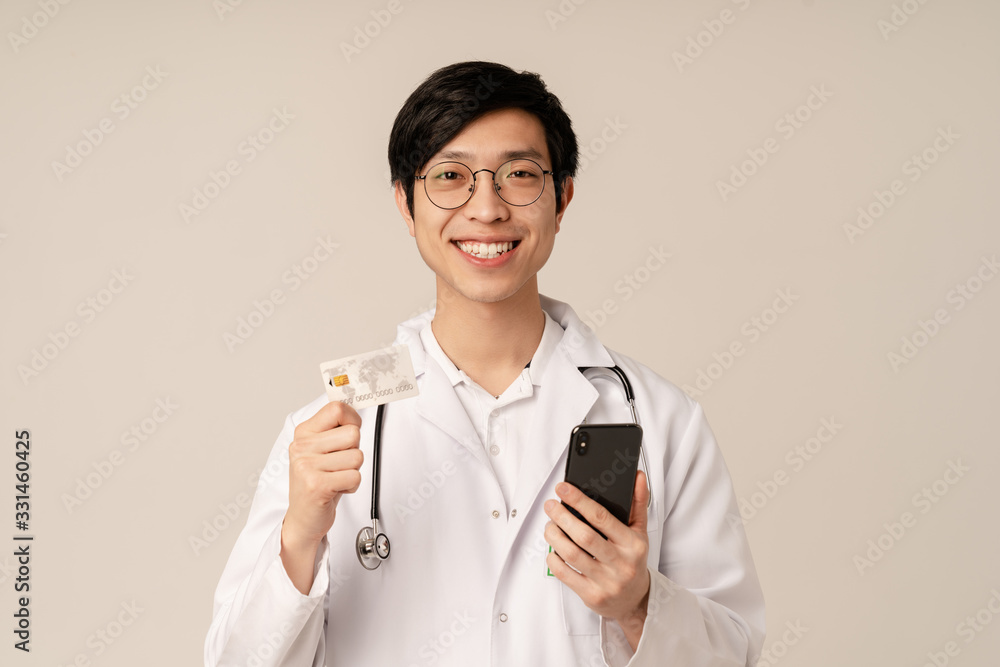 Image of asian young doctor in uniform holding credit card and cellphone Image of asian young doctor in uniform holding credit card and cellphone