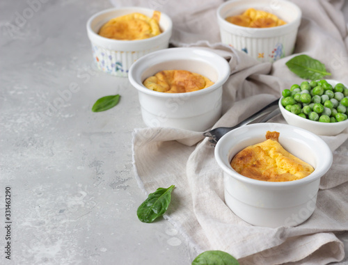 Cheese souffle with green peas in a white ceramic ramekin on a grey linen napkin. Light grey concrete background. 