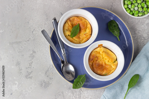 Two white ceramic ramekins with baked cheese souffle with green peas on a light grey table. Top view.