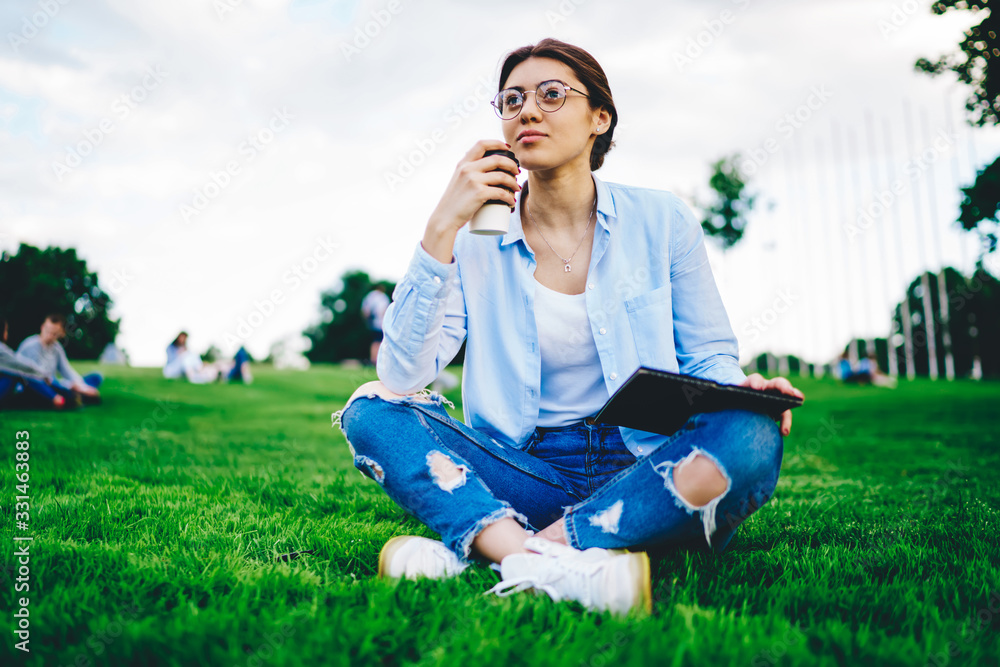 Pensive caucasian woman in casual wear and spectacles pondering on designing sketch holding coffee to go and resting on green grass, thoughtful hipster girl student recreating outdoors in campus Pensive caucasian woman in casual wear and spectacles pondering on designing sketch holding coffee to go and resting on green grass, thoughtful hipster girl student recreating outdoors in campus