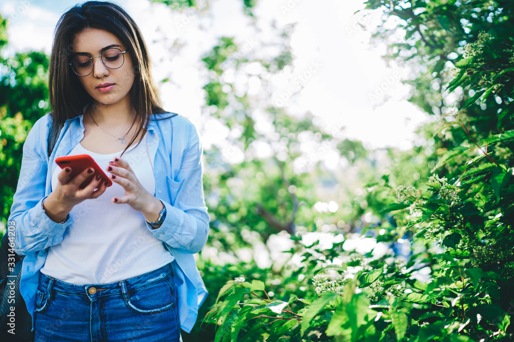 Pensive caucasian millennial woman standing on green trees background in park holding mobile phone, young brunette female blogger checking mail spending free time on sunny day outdoors Pensive caucasian millennial woman standing on green trees background in park holding mobile phone, young brunette female blogger checking mail spending free time on sunny day outdoors