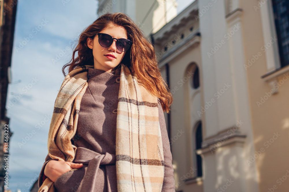 Street female fashion. Portrait of stylish young woman wearing coat glasses scarf on street outdoors. Spring accessories