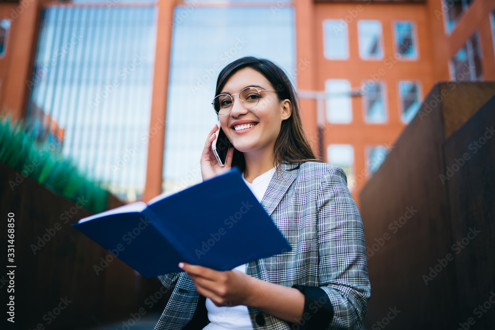Cheerful young female undergraduate talking on smartphone near college Cheerful young female undergraduate talking on smartphone near college