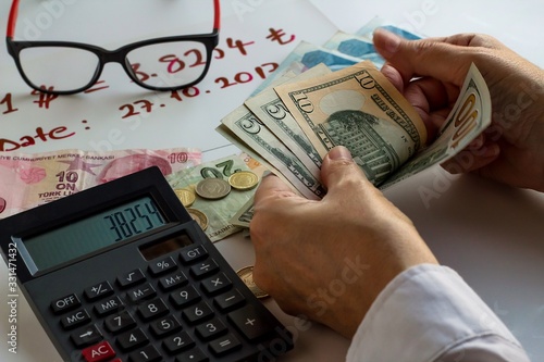 Fototapeta Naklejka Na Ścianę i Meble -  Hand counts dollars on white desk with calculator,glasses and Turkish Lira.Hand writing is over there exchange rate and date how much between dollar and lira.The rate also on calculator.
