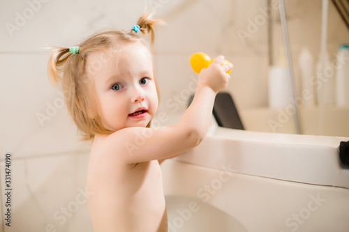 Toddler girl bathes in bath with foam at home, plays with rubber duck