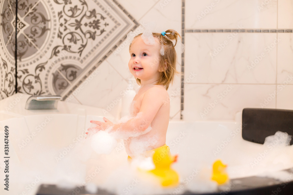 Toddler girl bathes in bath with foam at home, plays with rubber duck Toddler girl bathes in bath with foam at home, plays with rubber duck