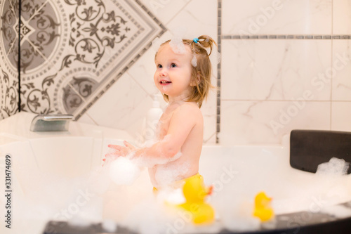 Toddler girl bathes in bath with foam at home, plays with rubber duck
