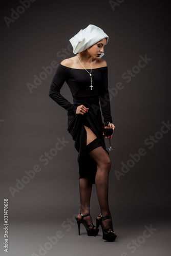 seductive nun in stockings holding glass of red wine on grey