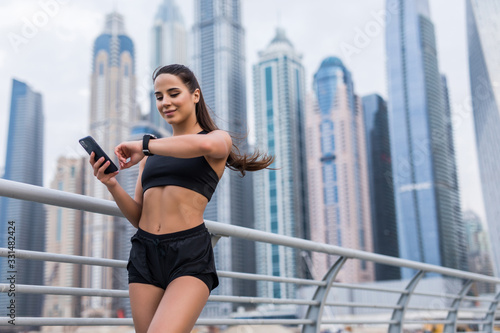 Photography Young woman holding phone with app for fitness smart watch after running on skyscrapers background