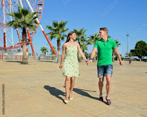 Romantic happy woman and man run. Palms tree and ferris wheel at background. Young couple walking outdoors in city. Hug near grey wall.