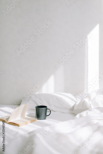 In the bright bedroom on the bed is an open old book next to a cup of coffee or tea. Against the background of the sun's rays. Vertical photo