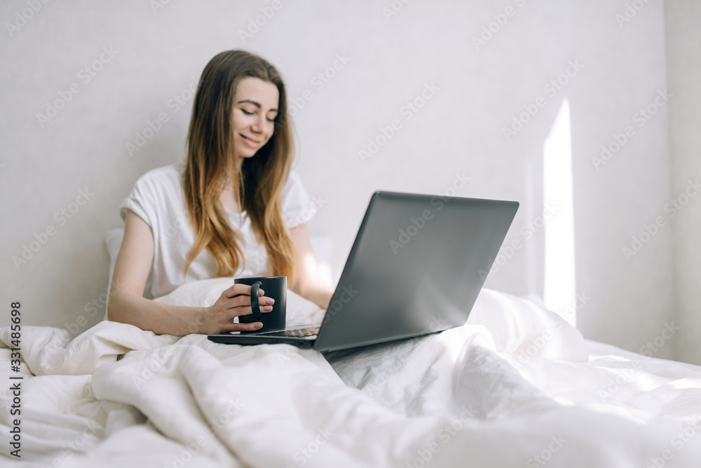 Beautiful woman in casual clothes working in the morning in bed on a laptop with a cup of coffee. Beautiful woman in casual clothes working in the morning in bed on a laptop with a cup of coffee.