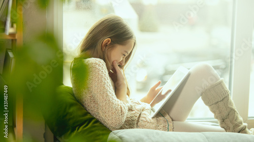 Little girl sitting near by window with tablet, remote studying, being at home. Doing homework, online serfing, chatting, conference with teacher. Staying home, indoors lifestyle, education concept.