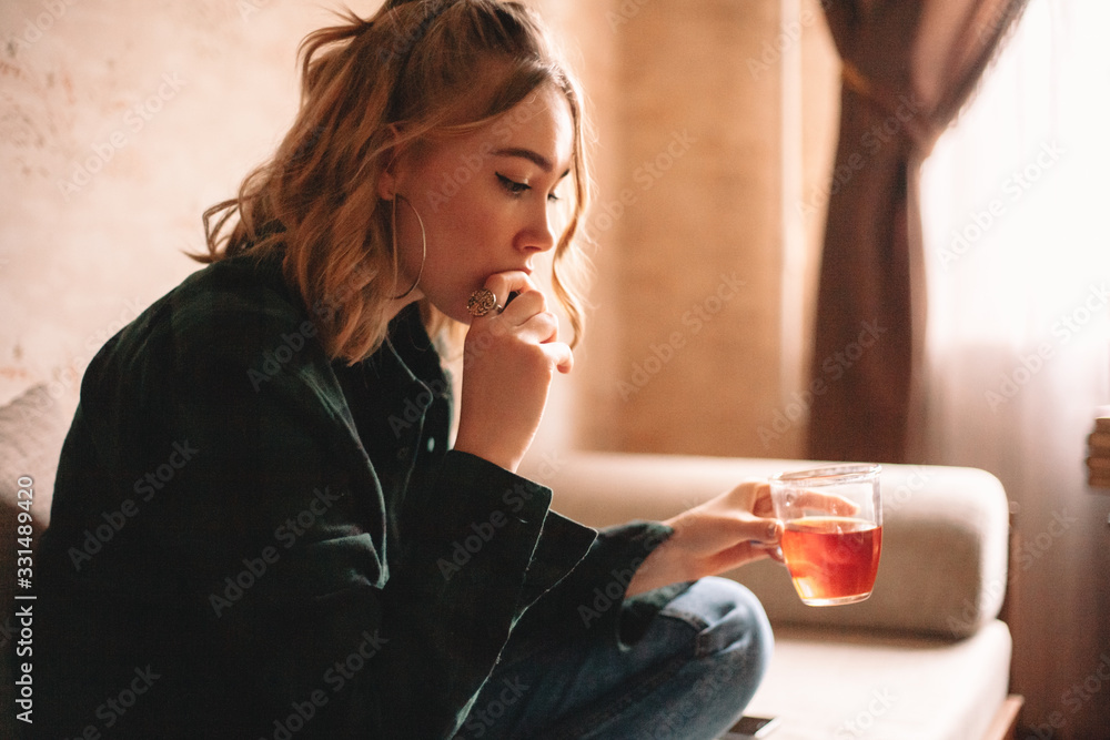 Naklejka premium Young thoughtful upset woman drinking tea and eating chocolate while sitting on sofa in living room at home