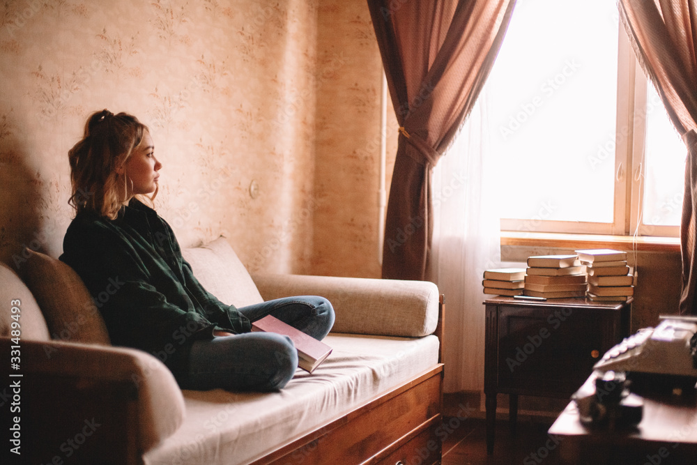 Young sad woman holding book looking through window while sitting on ...