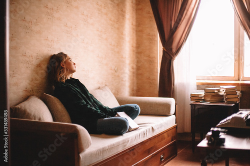 Young upset woman holding book looking through window while sitting on sofa in living room at home