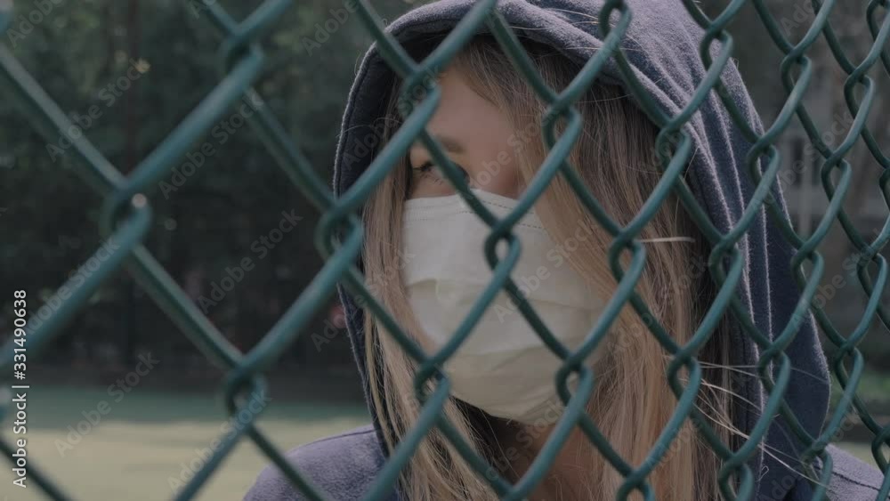 Closed border. Quarantine. Close-up of young woman wearing Protective Face Mask stand behind fence border. COVID-19 coronavirus infection pandemic disease virus. Closed border. Quarantine. Close-up of young woman wearing Protective Face Mask stand behind fence border. COVID-19 coronavirus infection pandemic disease virus.