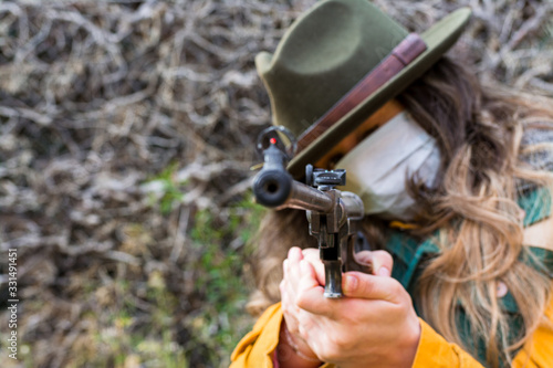 Blonde scout girl with mask, green vest and green hat aiming rifle at camera. Prepared to face the coronavirus.