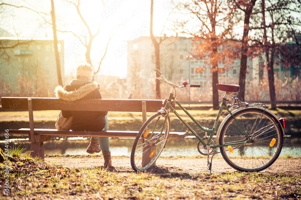 Enjoying the sun in spring: Young girl is sitting on park bench, bicycle back view