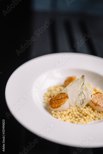 a plate with ptitim pasta  grilled scallops  cream sauce and dried onion slices. Dark background. Smooth image with shallow depth of field.