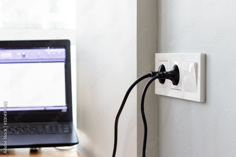 Group of white european electrical outlets with a two black plugs inserted into it and a switch located on a gray wall in a bright room with a blurred working laptop by the window. Selective focus Group of white european electrical outlets with a two black plugs inserted into it and a switch located on a gray wall in a bright room with a blurred working laptop by the window. Selective focus