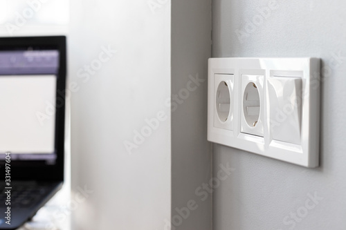 Group of white european electrical outlets and a switch located on a gray wall in a bright modern room with a blurred working laptop by the window. Selective focus. Closeup view