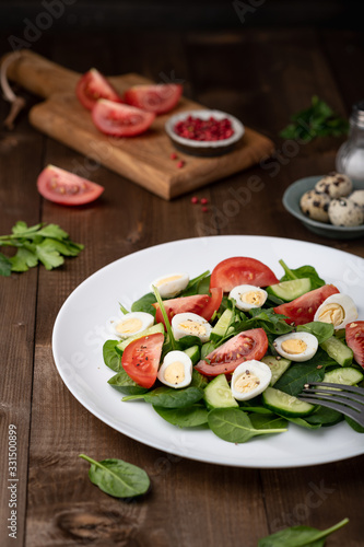 Salad with spinach, cucumber, tomato and quail egg on a white plate on a wooden background.