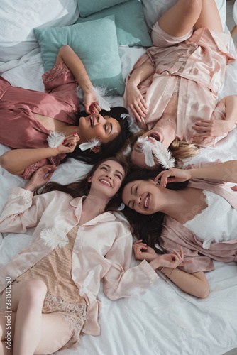 Top view of happy and excited multiethnic women resting on bed with feathers at bachelorette party