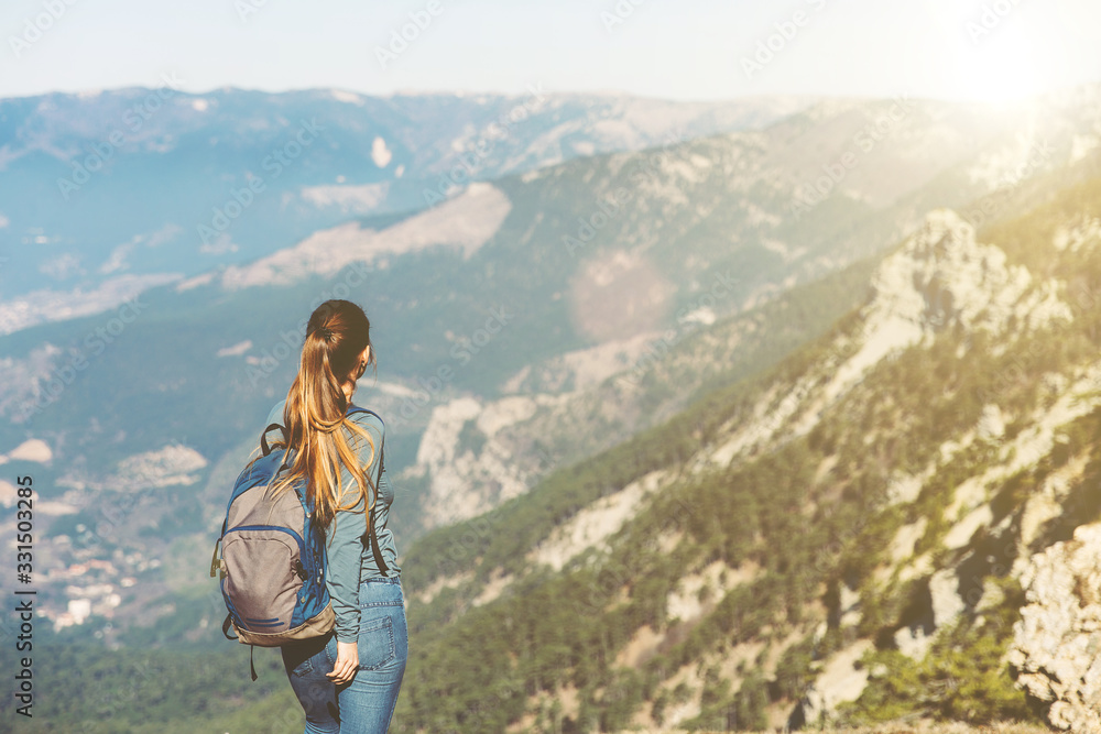 Obraz premium Young beautiful girl travels alone in the mountains in spring or autumn, looks into the distance and enjoys nature, rocks and green forests, view of the landscape. a backpack behind and sportswear