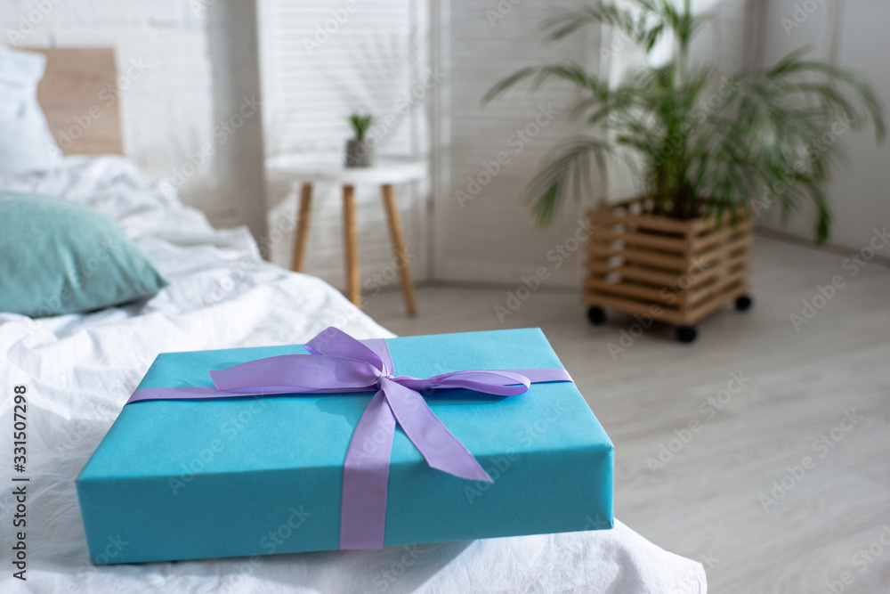 Selective focus of blue gift box with violet ribbon on bed in room Selective focus of blue gift box with violet ribbon on bed in room