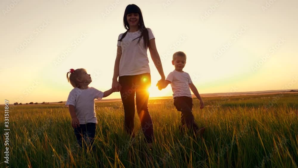 A young woman mother walks with two young children on the grass in the Park. A happy family walks outdoors in the setting sun. A young woman mother walks with two young children on the grass in the Park. A happy family walks outdoors in the setting sun.
