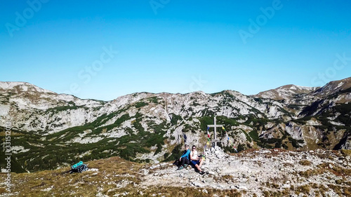 A drone shot of a couple in hiking outfits sitting next to a cross on top of Buchbergkogel, Hochschwab region, Austria. They are surrounded by mountains. Achieving a goal. Alpine landscape.