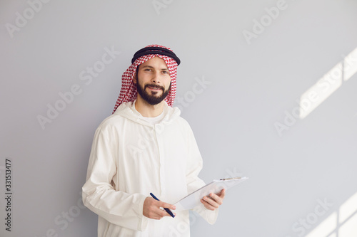 Tablou canvas Attractive smiling arab man writes in clipboard on gray background