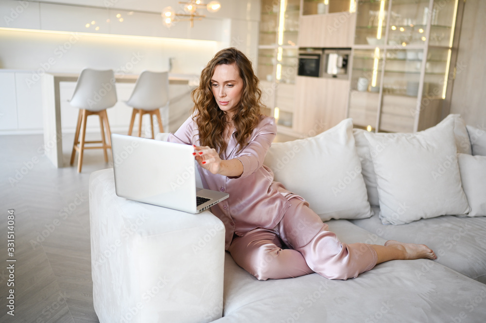 Young woman sitting on a white sofa in scandinavian style living room kitchen interior and working on a laptop. Home quarantine during Covid-19 pandemic Corona virus. Distance work from home concept. Young woman sitting on a white sofa in scandinavian style living room kitchen interior and working on a laptop. Home quarantine during Covid-19 pandemic Corona virus. Distance work from home concept.