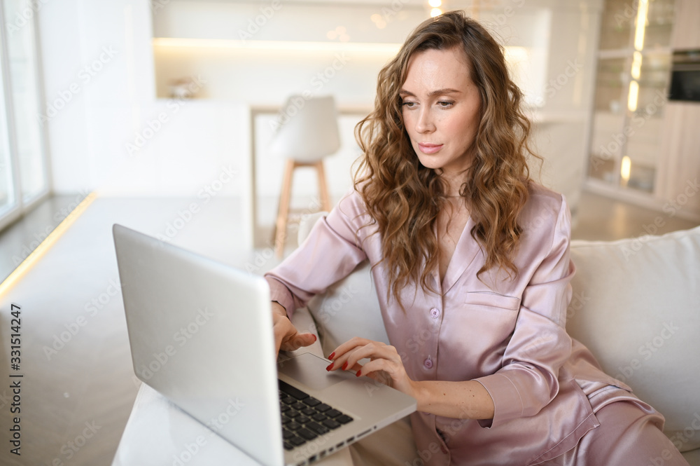 Young woman sitting on a white sofa in scandinavian style living room kitchen interior and working on a laptop. Home quarantine during Covid-19 pandemic Corona virus. Distance work from home concept. Young woman sitting on a white sofa in scandinavian style living room kitchen interior and working on a laptop. Home quarantine during Covid-19 pandemic Corona virus. Distance work from home concept.