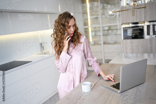 Young woman speaking smartphone and working on a laptop while drinking morning coffee. Home quarantine during Covid-19 pandemic Corona virus. Distance work from home concept. Modern kitchen interior